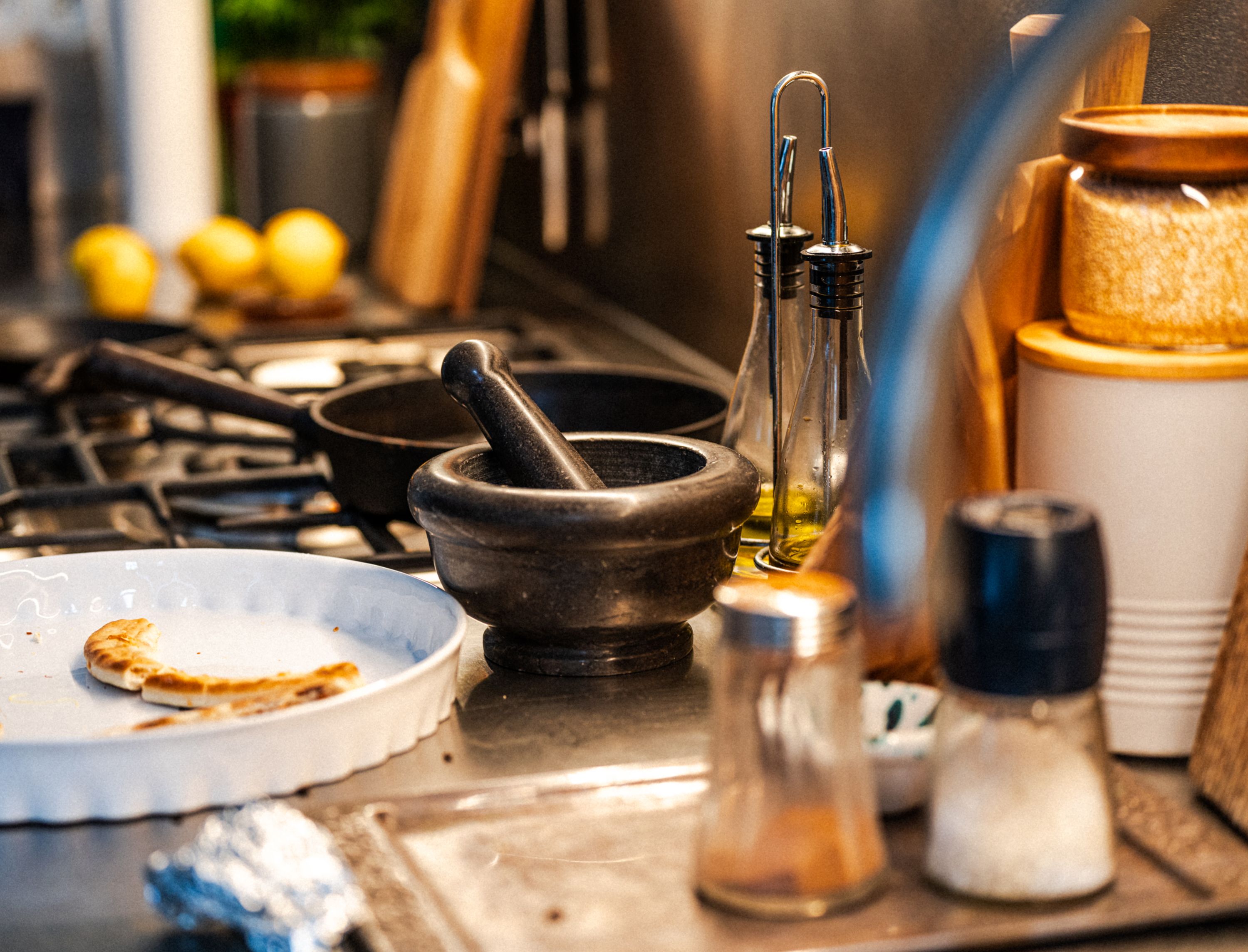 A mortar and pestle, olive oil, lemons and jars on a stovetop, the raw ingredients a recipe turns into a costed dish.