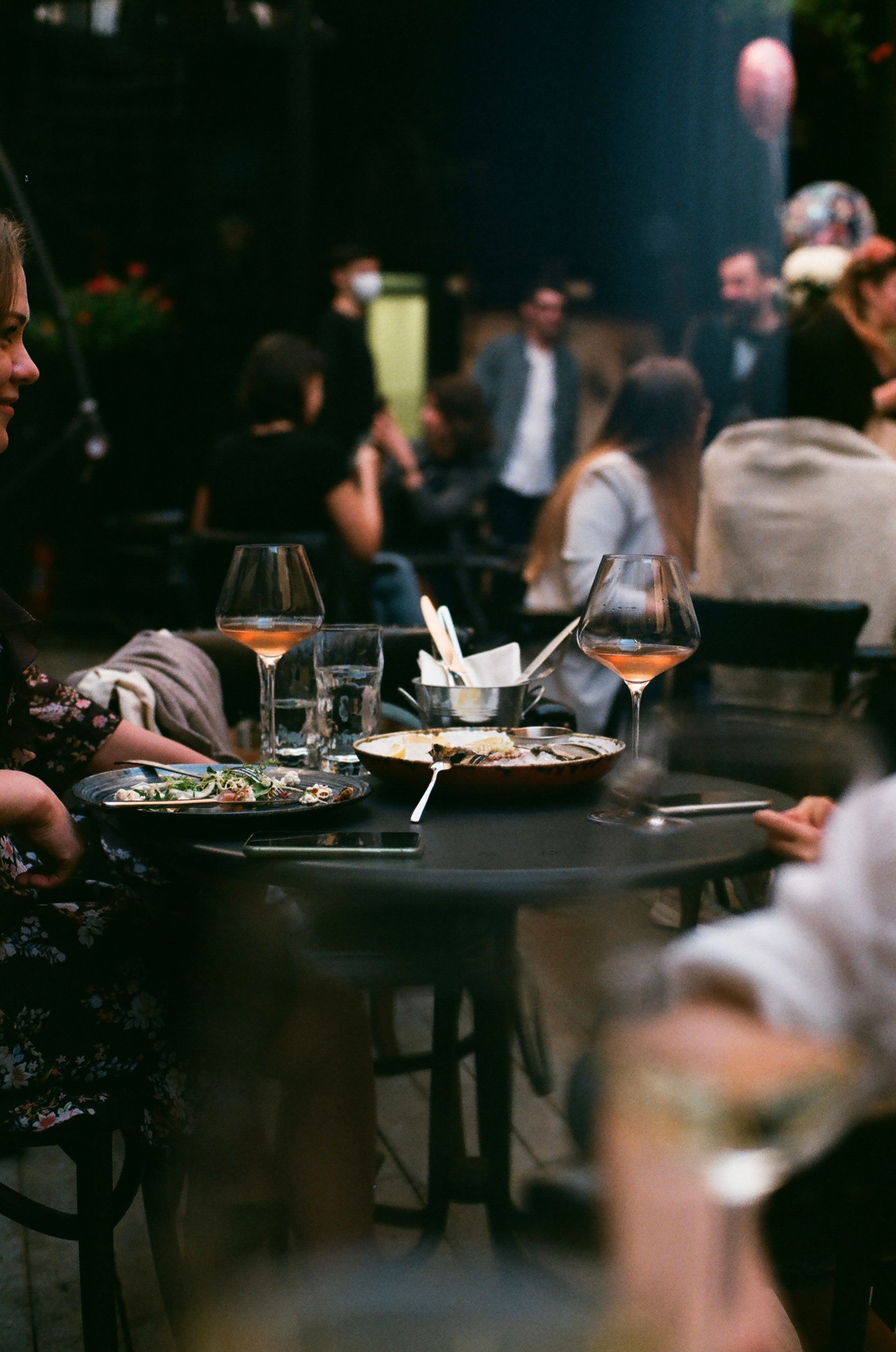 Guests at a small round table on a busy terrace in the evening, with glasses of wine and plates on the table.